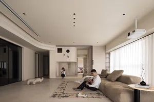 A modern minimalist living room with neutral-toned walls, furniture, and flooring, featuring a large sofa, a reading woman, a
