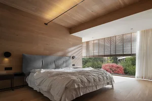 Modern bedroom with wooden walls, ceiling, and floor, featuring a comfortable bed with a grey cushioned headboard and neutral