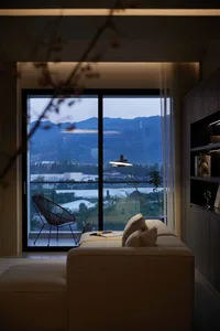 A modern minimalist living room with a view of distant mountains, featuring a white sofa, a metal chair, and a lamp on a soft