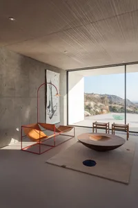 A spacious, minimalist living room featuring a concrete wall, wooden coffee table, and a large window offering a scenic view.