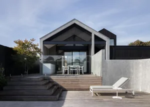 A modern house with a concrete exterior, glass windows, and wooden stairs leading to a wooden terrace with a table and chairs