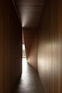 A narrow hallway with wooden walls, floor, and ceiling, featuring a window at the end.