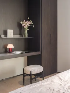 A minimalist bedroom featuring wooden furniture, a bed, a desk with a lamp and flowers, and a shelf.