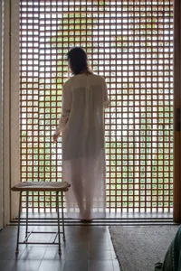 A woman is standing in front of a wooden lattice window with a stool in the foreground and a textured rug on the floor.