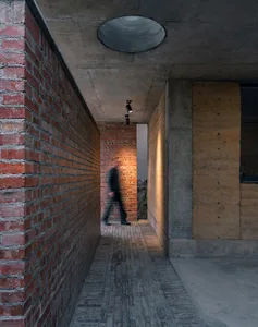 A minimalist corridor with brick and concrete walls, a concrete ceiling, and a person walking through a doorway.