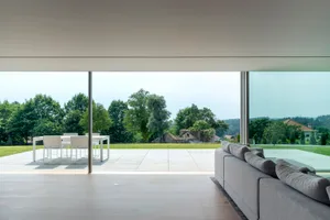 Modern minimalist living room with a large glass wall overlooking a lush green landscape.