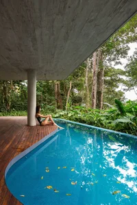 A woman sitting by a modern, concrete-covered pool surrounded by lush tropical vegetation.