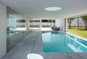 Modern residential swimming pool with clear glass walls and a view of the outdoor area with greenery and a round sofa.