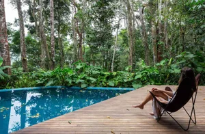 Person lounging on wooden deck overlooking lush green poolside retreat surrounded by plants.