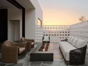 Modern outdoor lounge area with concrete and rattan furniture, glass fire pit, and sunset view.