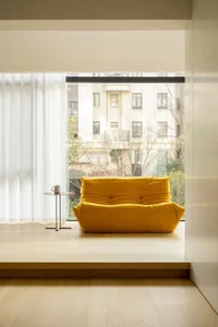 A minimalist yellow sofa sits in front of a window, with a small table beside it. The window has a white curtain, and the bac