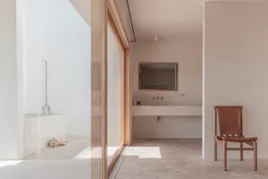 A minimalist bathroom with a concrete floor and bathtub, featuring a wooden chair and a concrete sink with a mirror and fauce