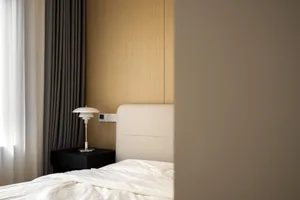 A minimalist bedroom with a white bed, grey curtains, a black side table with a white lamp, and a wooden headboard.