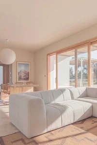 Minimalist living room with a white couch, dining table, and large windows allowing natural light to enter.