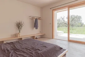 A minimalist modern bedroom with a concrete floor and wall, featuring a bed with a gray blanket and a small plant.