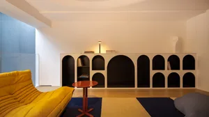 A modern living room featuring a yellow couch, a wooden side table, a bookshelf with arched openings, and a dark wooden floor