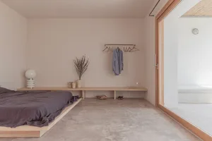 Spacious minimalist bedroom with concrete floor and wall, featuring a low-profile bed, modern lamp, and natural light.