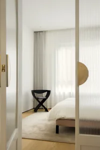 A minimalist bedroom with white curtains, a white bedspread, and a black chair.