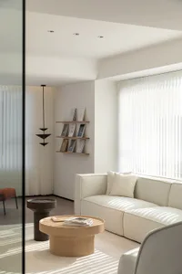A minimalist living room featuring a light wood sofa and coffee table, with white curtains and a suspended light fixture.