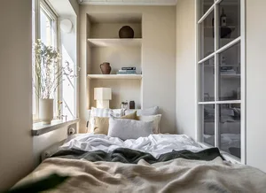 A cozy, minimalist bedroom with neutral-colored bedding, shelves filled with books and vases, and a window letting in natural