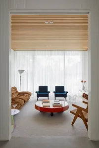 A modern living room with transparent windows, featuring two blue armchairs, a brown sofa, a round coffee table, and a white 