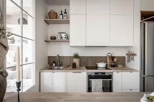 A brightly lit, modern kitchen with light wood countertops and minimalist white cabinetry, featuring a sink, stove, and oven.