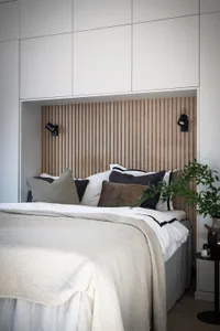 Modern minimalist bedroom interior with a wooden paneled headboard, white bedding, and a green plant on a side table.