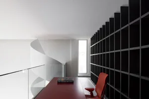 A modern minimalist office library with a red table, bookcase, and spiral staircase.