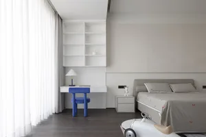 Modern, minimalist bedroom featuring a white bed with neutral bedding, a blue chair, and white shelves with a lamp on a desk.