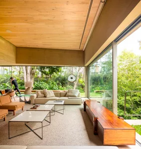A minimalist living room with wooden furniture and glass walls, featuring a view of the greenery outside.