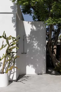 A minimalist outdoor shower with a concrete wall and tree shadows.