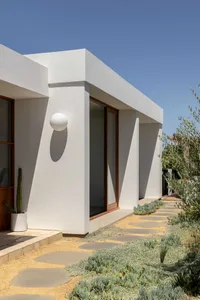 A modern residential exterior with a white plaster facade, sand pathway, and green plants