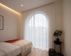A minimalist white bedroom with a large arched window covered in white curtains, a white bed with white sheets and a white bl