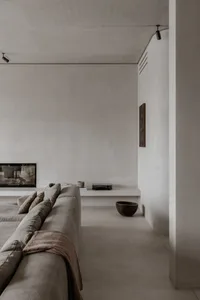 A minimalist living room with a concrete floor, a large grey sofa, and a built-in entertainment unit with a television and a 