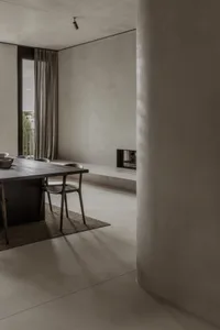 A modern living room featuring a minimalist concrete fireplace, wooden table, and area rug, with a large window providing nat