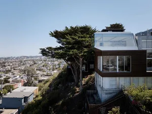 A modern residential building with concrete exterior, set on a hill overlooking a cityscape.