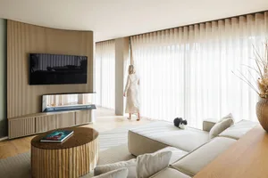 A modern living room with neutral tones and a cat resting on a sofa, as a woman stands by sheer curtains, looking out large w