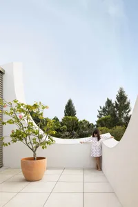 A modern rooftop garden featuring a child leaning against a curved white wall on a concrete floor, surrounded by greenery.