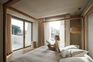 A modern minimalist bedroom with natural wooden elements, featuring a woman reading a book seated on the bed, overlooking a b