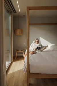A woman in a minimalist bedroom reading a book while sitting on a wooden canopy bed with white linens and cushions, illuminat