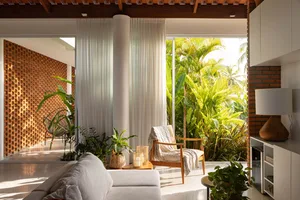 A minimalist living room with white curtains, a white fabric sofa, and wooden armchair surrounded by green plants.