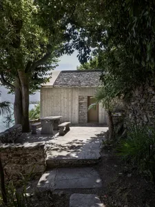 A minimalist house with a stone exterior surrounded by lush greenery, featuring a stone pathway and a wooden cabin.