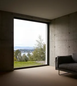 Modern, minimalist living room with a concrete wall and large window overlooking green hills and water.