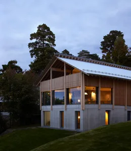 A modern building with a concrete base and wooden lattice facade standing on a grassy hill with tall trees in the background.