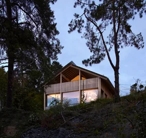 A modern residential house with large windows and a wooden exterior, situated on a grassy hillside with trees.