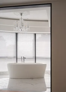 A modern bathroom with a freestanding bathtub made of white marble, featuring a crystal chandelier and blinds in front of the