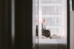 A woman with pastel-colored hair sits cross-legged on a window sill in a minimalist interior, reading a book with a glass of 