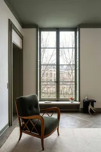 A green armchair with a wooden frame in a modern living room with large windows.