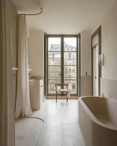 A modern minimalist bathroom with a concrete bathtub and flooring, large windows with a view of the city, and a simple chair.
