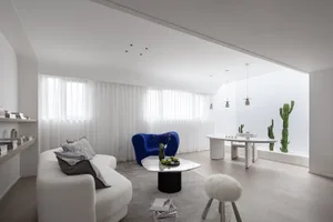 A modern minimalist living room featuring a white couch, blue accent chair, and glass coffee table with a vase and green appl